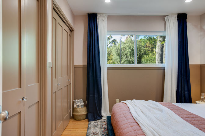 Cozy residential bedroom with blush quilt and white throw on bed, navy and white curtains framing a sunlit window with green tree view, paneled tan closet doors, wicker basket, hardwood floor and patterned rug.