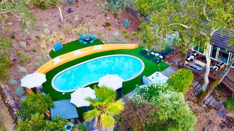 Aerial view of a kidney-shaped backyard pool on green turf with white umbrellas and loungers, adjacent wooden deck with outdoor seating and hot tub, palm tree and dry hillside landscaping.