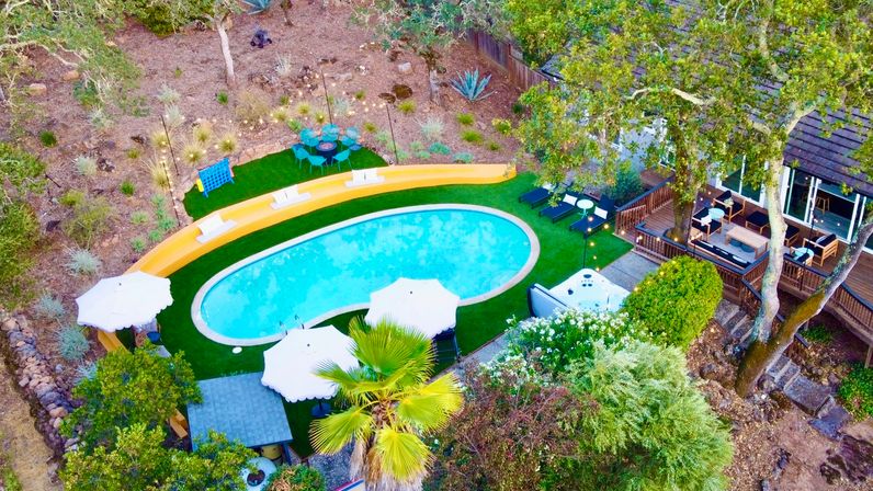 Aerial view of a kidney-shaped backyard pool on green turf with white umbrellas and loungers, adjacent wooden deck with outdoor seating and hot tub, palm tree and dry hillside landscaping.