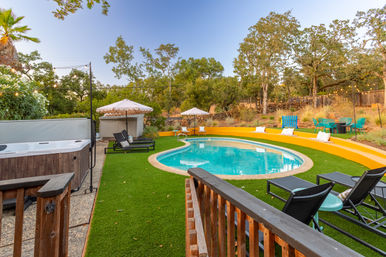 Backyard curved swimming pool with turquoise water, surrounded by artificial turf, a hot tub, black lounge chairs, scalloped umbrellas, a bright yellow curved seating wall, string lights and oak trees on a wooded hillside.
