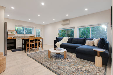 Bright open-plan living room and kitchen with a navy L-shaped sofa, round wooden coffee table, patterned area rug, island with three wood stools, and large leafy windows.