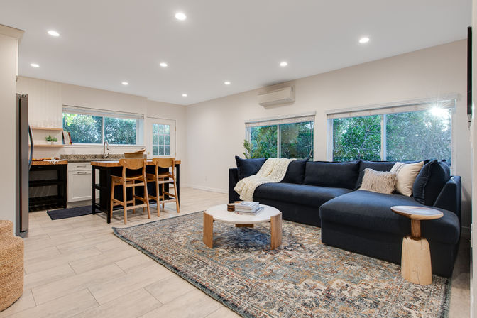 Bright open-plan living room and kitchen with a navy L-shaped sofa, round wooden coffee table, patterned area rug, island with three wood stools, and large leafy windows.