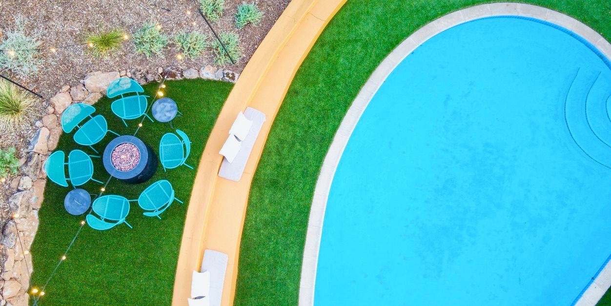 Aerial view of a curving backyard pool with bright turquoise water, adjacent artificial turf patio featuring a circular cluster of teal chairs around a fire pit, mustard-yellow curved seating wall with white cushions and string lights.