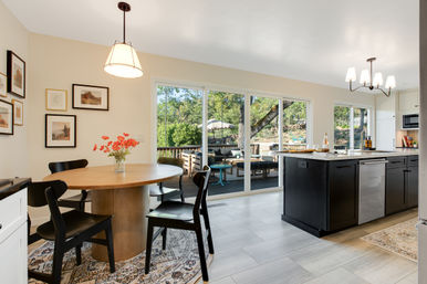 Sunny open-concept kitchen and dining area with round wooden table and black chairs, vase of flowers, dark island with marble countertop and dishwasher, and large sliding glass doors opening to a shaded backyard deck with outdoor seating and umbrella.