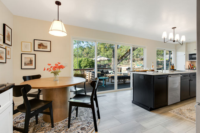 Sunny open-concept kitchen and dining area with round wooden table and black chairs, vase of flowers, dark island with marble countertop and dishwasher, and large sliding glass doors opening to a shaded backyard deck with outdoor seating and umbrella.