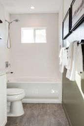 Bright modern white bathroom with bathtub, handheld showerhead, toilet, window, hanging white towels, textured wall tile and gray wood-look floor