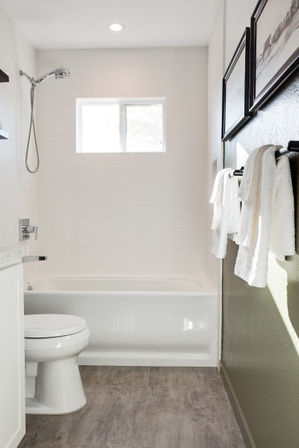 Bright modern white bathroom with bathtub, handheld showerhead, toilet, window, hanging white towels, textured wall tile and gray wood-look floor