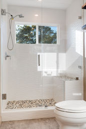 Bright modern bathroom with glass-enclosed walk-in shower, white textured tile walls, pebble mosaic shower floor, built-in bench, handheld showerhead, small window with tree view and white toilet in foreground.