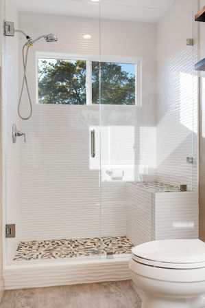 Bright modern bathroom with glass-enclosed walk-in shower, white textured tile walls, pebble mosaic shower floor, built-in bench, handheld showerhead, small window with tree view and white toilet in foreground.