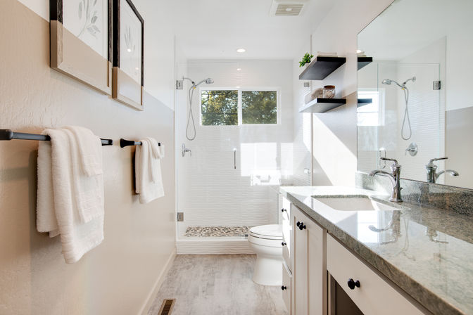 Bright modern residential bathroom with glass walk-in shower, granite vanity with chrome faucets, white towels on a dark towel bar, floating shelves and natural light