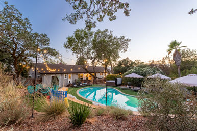 Twilight suburban backyard oasis with a curved swimming pool reflecting string lights, lounge chairs and umbrellas, a hot tub by a cozy single-story house framed by oak and palm trees.