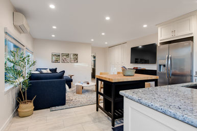 Bright modern open-concept living room and kitchen with a navy sofa, potted plant by the window, wood-topped island, granite countertops, stainless steel refrigerator, wall-mounted TV, and recessed lighting.
