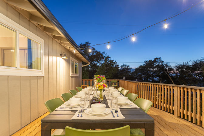 Twilight outdoor dining on a wooden home deck with a long table set for guests, green chairs, string lights and a floral centerpiece overlooking treetops.