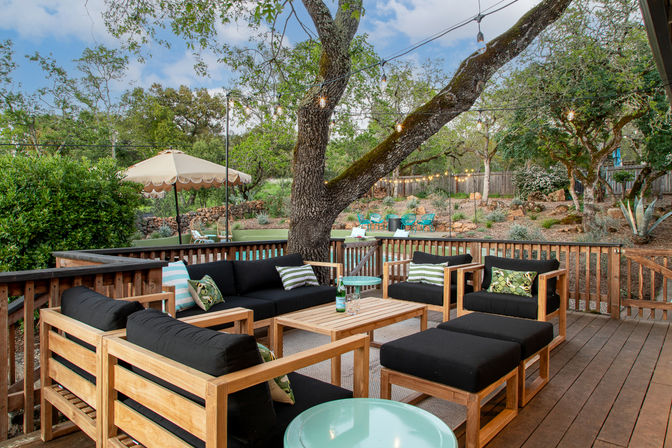 Cozy backyard wooden deck with teak-style outdoor sofa and armchairs with black cushions, string lights draped from a large oak tree and a landscaped rock garden