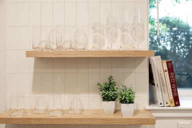 Sunlit modern kitchen with wooden floating shelves displaying clear drinking and wine glasses, two small potted green plants, and a stack of cookbooks by a window against a white tile backsplash.