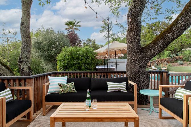 Sunny backyard deck with wooden outdoor sofa set and black cushions, striped throw pillows, a wooden coffee table holding a bottle of sparkling water and glass, large oak trees with string lights, scalloped patio umbrella and a pool surrounded by lush greenery.