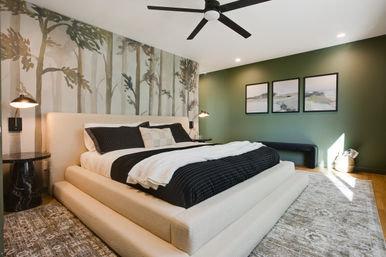 Cozy modern master bedroom with plush cream platform king bed, black-and-white bedding, forest mural accent wall, green painted wall with three landscape prints, black ceiling fan, bedside lamps, and textured area rug.