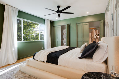 Sunlit modern bedroom with sage-green walls, large window with white curtains, cream upholstered platform bed dressed in white linens and a black throw, black ceiling fan, and built-in closets with open shelving.