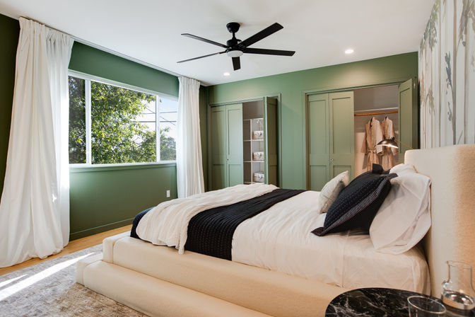 Sunlit modern bedroom with sage-green walls, large window with white curtains, cream upholstered platform bed dressed in white linens and a black throw, black ceiling fan, and built-in closets with open shelving.
