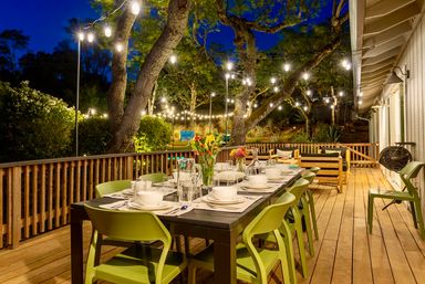 Cozy twilight backyard wooden deck with a long outdoor dining table set for dinner, lime-green chairs, and string lights draped among mature trees.