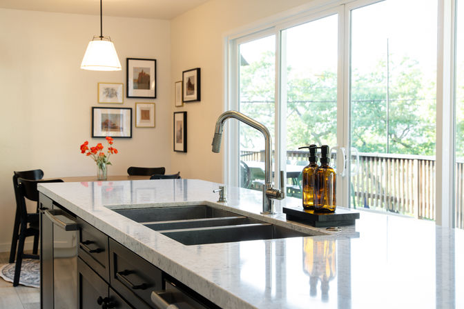 Sunlit open-plan kitchen island with white marble countertop, double stainless sink and gooseneck faucet, amber soap dispensers on a tray, vase of orange flowers on a nearby dining table, framed wall art and sliding glass doors overlooking a leafy backyard deck.