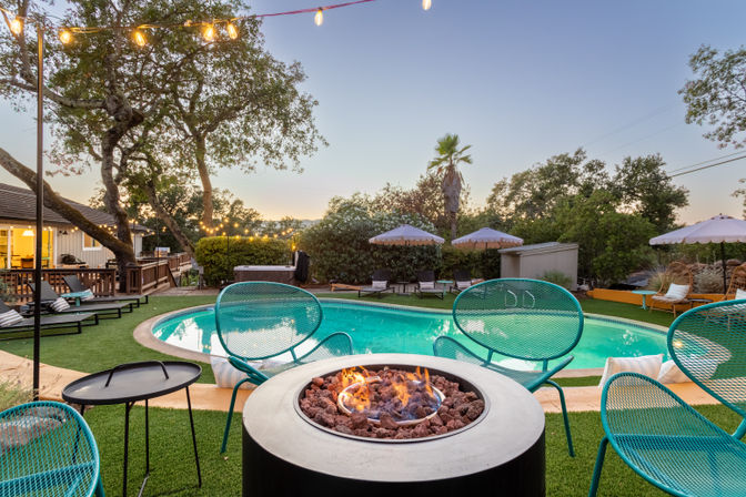 Backyard pool at dusk with a glowing round fire pit surrounded by teal metal chairs, string lights overhead, turquoise swimming pool, lounge chairs and umbrellas on artificial turf.