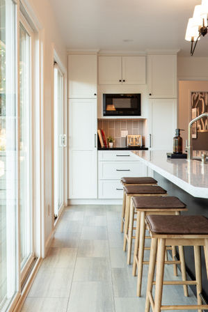 Sunlit modern white kitchen with light stone island and four wooden bar stools, sliding glass doors, built-in microwave, toaster and white shaker cabinets
