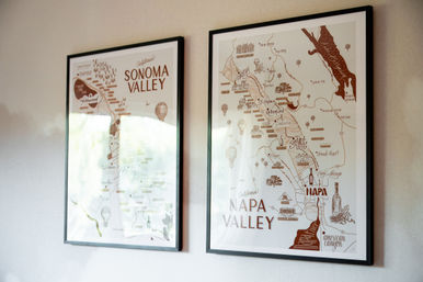 Two framed illustrated maps of California wine regions — Sonoma Valley and Napa Valley — hung on a light wall, warm vintage-style artwork with soft glass reflections.