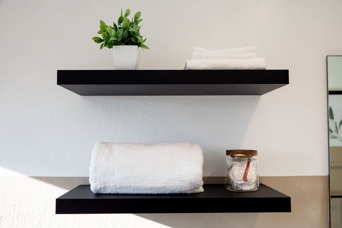 Minimalist home bathroom decor: black floating shelves on a textured white wall holding a potted green plant, folded white towels and a glass jar with cotton pads.