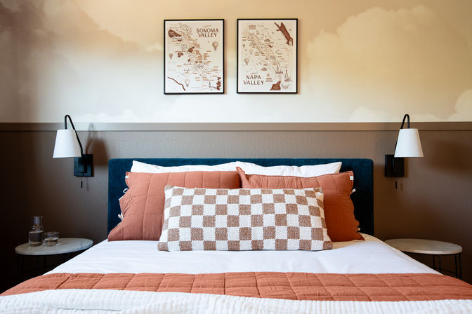 Cozy modern bedroom with teal upholstered headboard, rust-orange quilt and pillows, checkered lumbar cushion, framed Napa Valley and Sonoma Valley map prints above the bed, wall sconces and round marble bedside tables.
