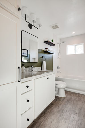 Bright modern bathroom with white vanity and marble countertop, black-framed mirror and matte-black hardware, floating shelves, bathtub with handheld shower, and wood-look tile floor.