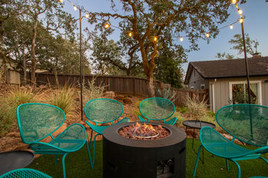Cozy suburban backyard patio at dusk with a round gas fire pit, teal mesh chairs clustered around it, string lights strung between oak trees, drought-tolerant plants and a small house in the background.