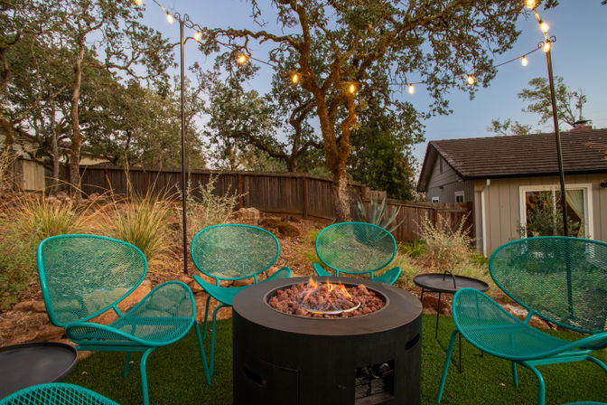 Cozy suburban backyard patio at dusk with a round gas fire pit, teal mesh chairs clustered around it, string lights strung between oak trees, drought-tolerant plants and a small house in the background.