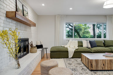 Cozy modern living room with green sectional sofa, chunky knit throw and striped pillows, white painted brick fireplace with wooden mantel, low wood coffee table, woven poufs, yellow flowering branches, and a large window framing leafy trees.
