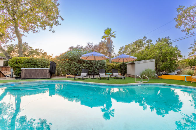 Suburban backyard pool at dusk with turquoise water reflecting a palm tree and sky, three lounge chairs under pastel umbrellas on a green lawn, hot tub and small shed framed by leafy trees.