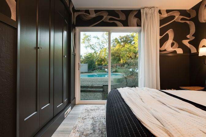Cozy modern bedroom with sliding glass door to a backyard pool, black built-in cabinets, abstract swirl wallpaper, and white quilted bedding.