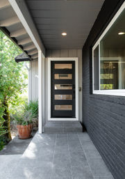 Sleek modern covered front porch with gray tile flooring, black door featuring five horizontal glass panels, black brick exterior wall, potted greenery and recessed ceiling light.