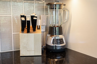 Wooden knife block with black-handled knives beside a stainless-steel blender on a glossy black granite kitchen counter with textured tile backsplash, ready for meal prep.