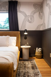 Cozy modern bedroom corner with tan upholstered bed and white linens, abstract ribbon-pattern wallpaper, black wall sconce over a round white bedside table with a glass carafe, woven basket of blankets on hardwood floor and a patterned rug.