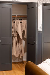 Cozy bedroom closet with two beige waffle-knit bathrobes hanging on a wooden rod behind dark gray doors, hardwood floor and bed corner visible.