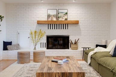 Cozy modern farmhouse living room with white painted brick fireplace and wooden mantel, framed landscape art, black fireplace insert, green sofa with knit throw, woven poufs, wood coffee table with books, yellow branch arrangement and a basket of firewood.