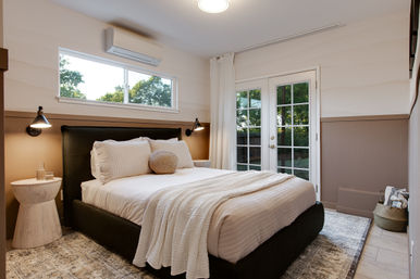 Cozy modern bedroom with queen bed, white linens and textured round pillow, black upholstered headboard, wall-mounted reading lamps, small wooden side table, soft area rug, and French doors plus high window letting in natural light and outdoor greenery.