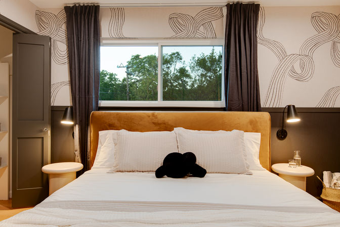 Cozy modern bedroom with a mustard velvet headboard, crisp white bedding and a black accent pillow, round bedside tables with wall lamps, patterned wallpaper and a window framing green trees outside.