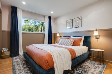 Bright cozy bedroom with large window and palm-tree view, navy curtains, blue upholstered bed with rust-orange quilt, checkered accent pillow, white throw, wall sconces and patterned rug.