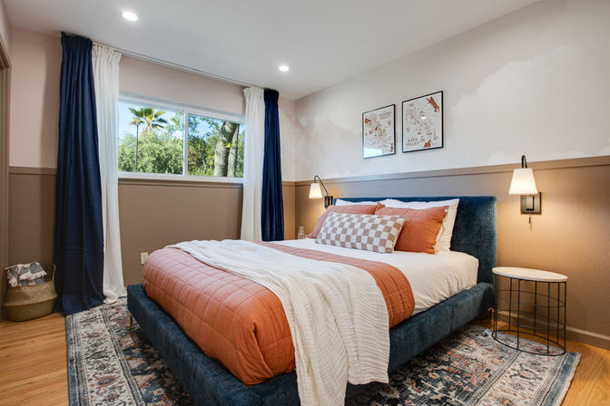 Bright cozy bedroom with large window and palm-tree view, navy curtains, blue upholstered bed with rust-orange quilt, checkered accent pillow, white throw, wall sconces and patterned rug.