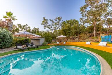 Curved turquoise backyard pool with concrete edge and artificial turf, bright yellow terraced seating with pillows, black lounge chairs under scalloped umbrellas, palm tree, hillside oaks, string lights and a giant outdoor Connect Four game.