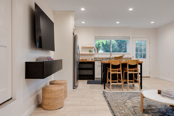 Bright modern open-plan kitchen and living area with a wooden island and three bar stools, stainless steel fridge, granite countertops, large window overlooking leafy greenery, wall-mounted TV, woven pouf stools and a textured area rug in neutral tones.
