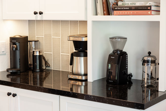Kitchen coffee station on black granite countertop with white cabinets — single-serve brewer, milk frother, stainless drip coffee pot, burr grinder, and French press beneath open shelves with cookbooks.