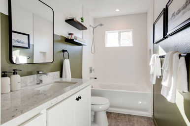 Bright contemporary bathroom with marble-look vanity and undermount sink, white bathtub, olive-green accent wall, floating shelves, small window and crisp white towels.