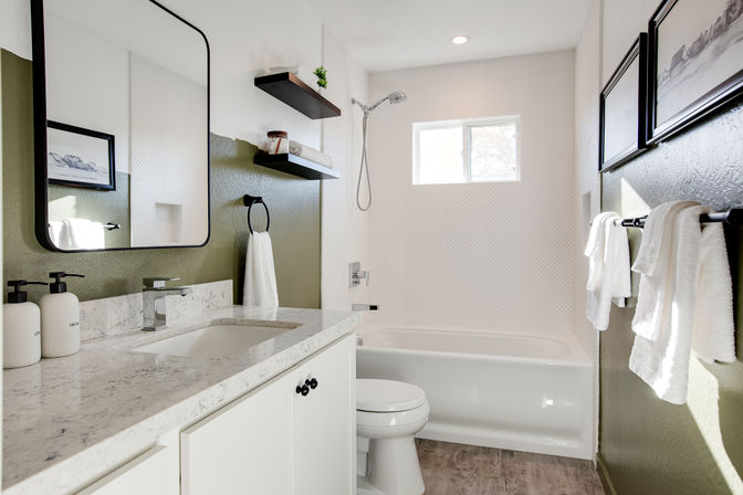 Bright contemporary bathroom with marble-look vanity and undermount sink, white bathtub, olive-green accent wall, floating shelves, small window and crisp white towels.